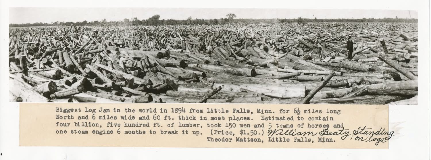 Panoramic photograph of a large logjam with workers standing among the logs.