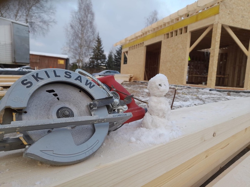 A circular saw and small snowman on snow-covered lumber, with a framed building behind them.